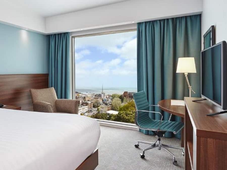 A hotel room with a large bed, a desk with a chair, a TV, and a view of the city and sea through a large window.