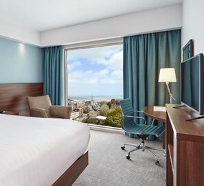 A hotel room with a large bed, a desk with a chair, a TV, and a view of the city and sea through a large window. - Hampton by Hilton Bournemouth (Bournemouth)