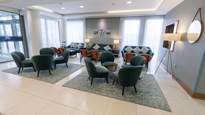 A modern hotel lobby with a seating area featuring green armchairs and a sofa set, a large window with a view, and a floor lamp.