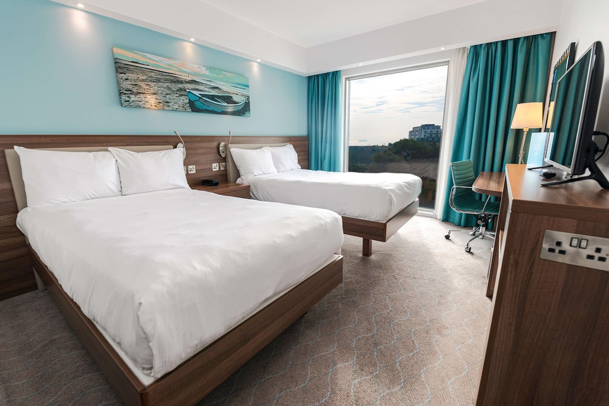 A hotel room with two beds, a desk with a computer, a chair, and a large window with curtains.