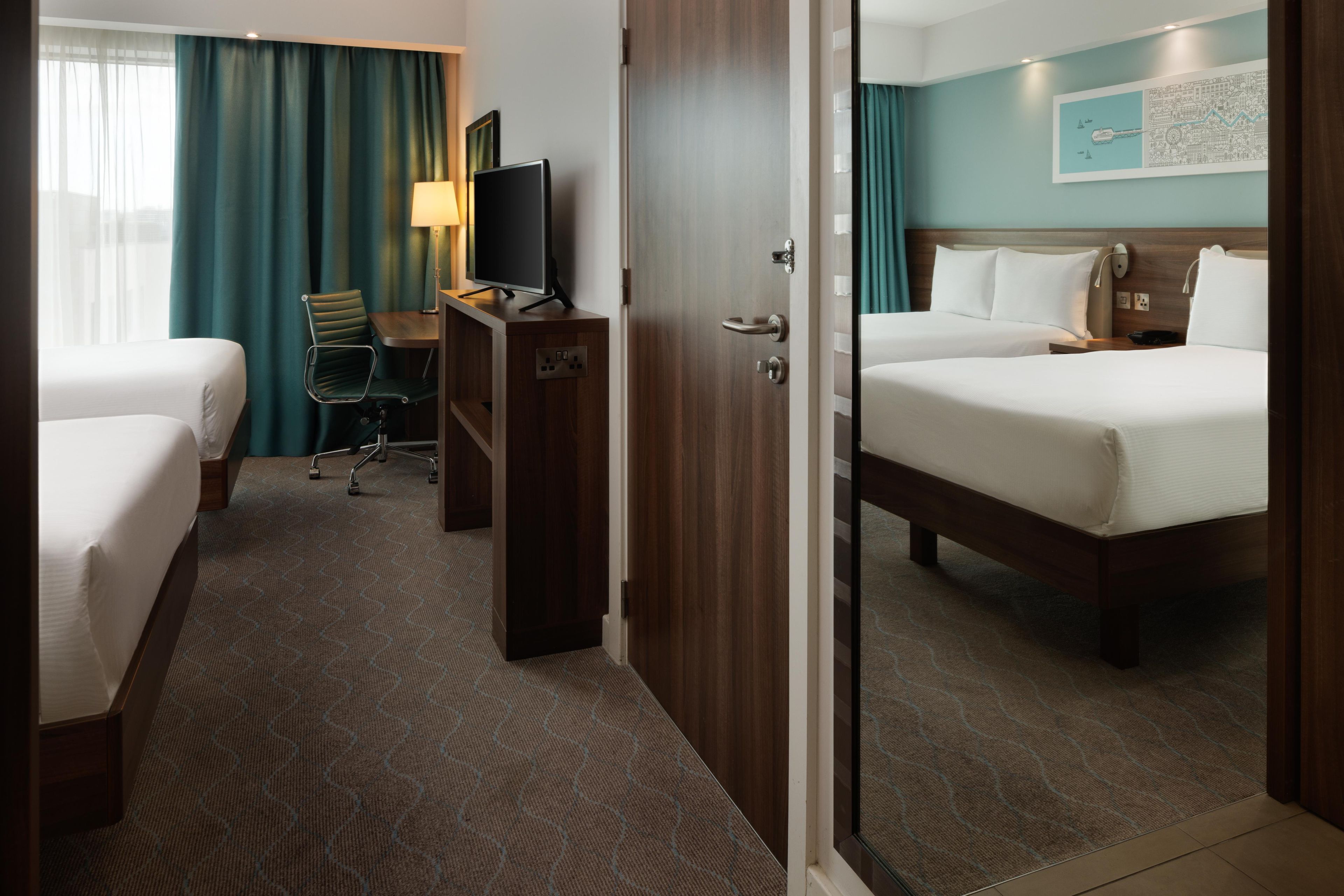 Ein Hotelzimmer mit zwei Betten, einem Schreibtisch mit Fernseher, einem Sessel und einer Tür zu einem weiteren Raum.