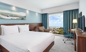 Habitación de hotel con una cama grande, un escritorio con televisor, una silla y vistas a la ciudad y al océano a través de un ventanal amplio.