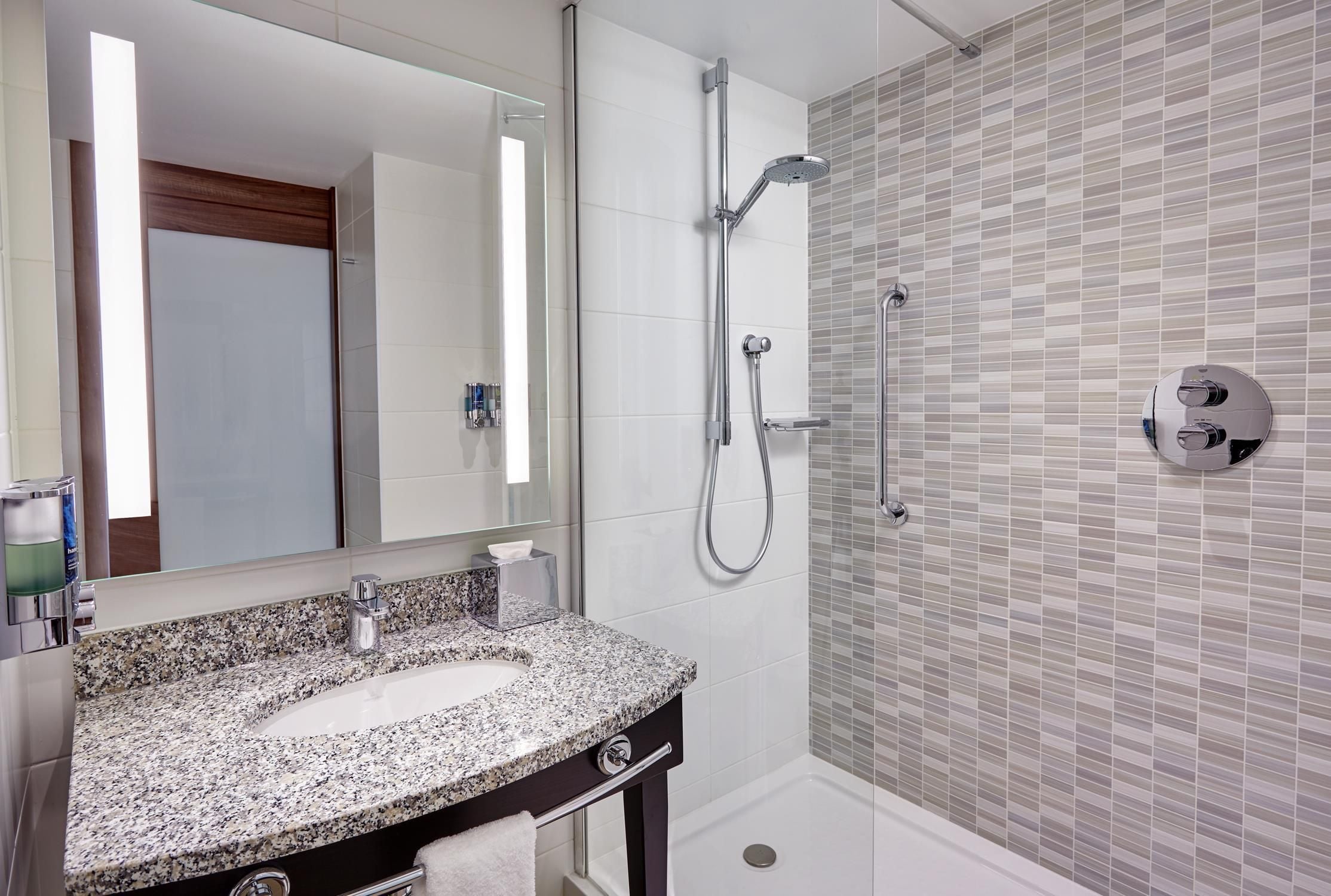 A modern bathroom with a granite countertop sink, a mirror, a shower with a handheld showerhead, and a tiled wall.