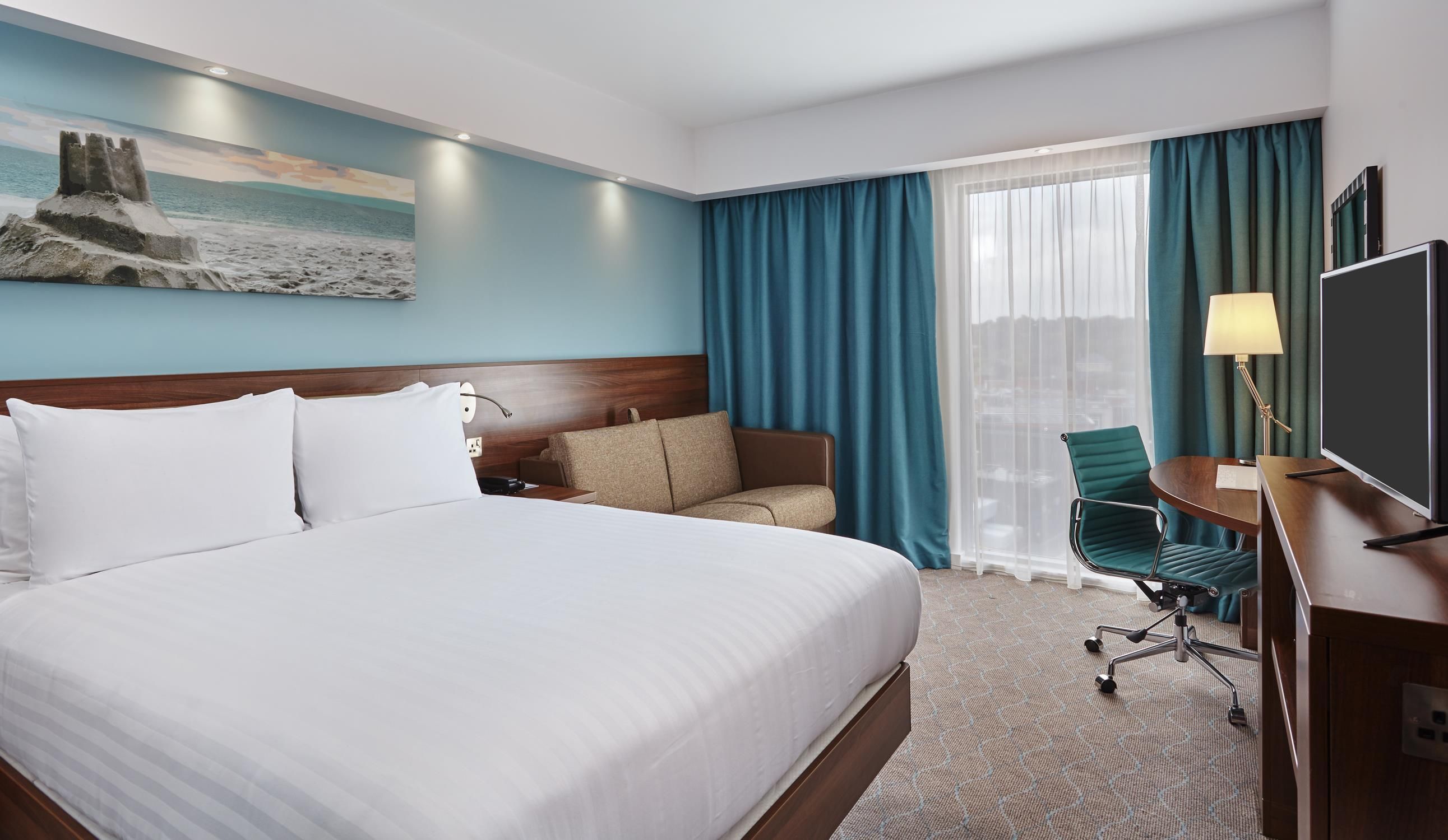 Habitación de hotel con una cama grande, un escritorio con silla, televisión y ventana con cortinas.
