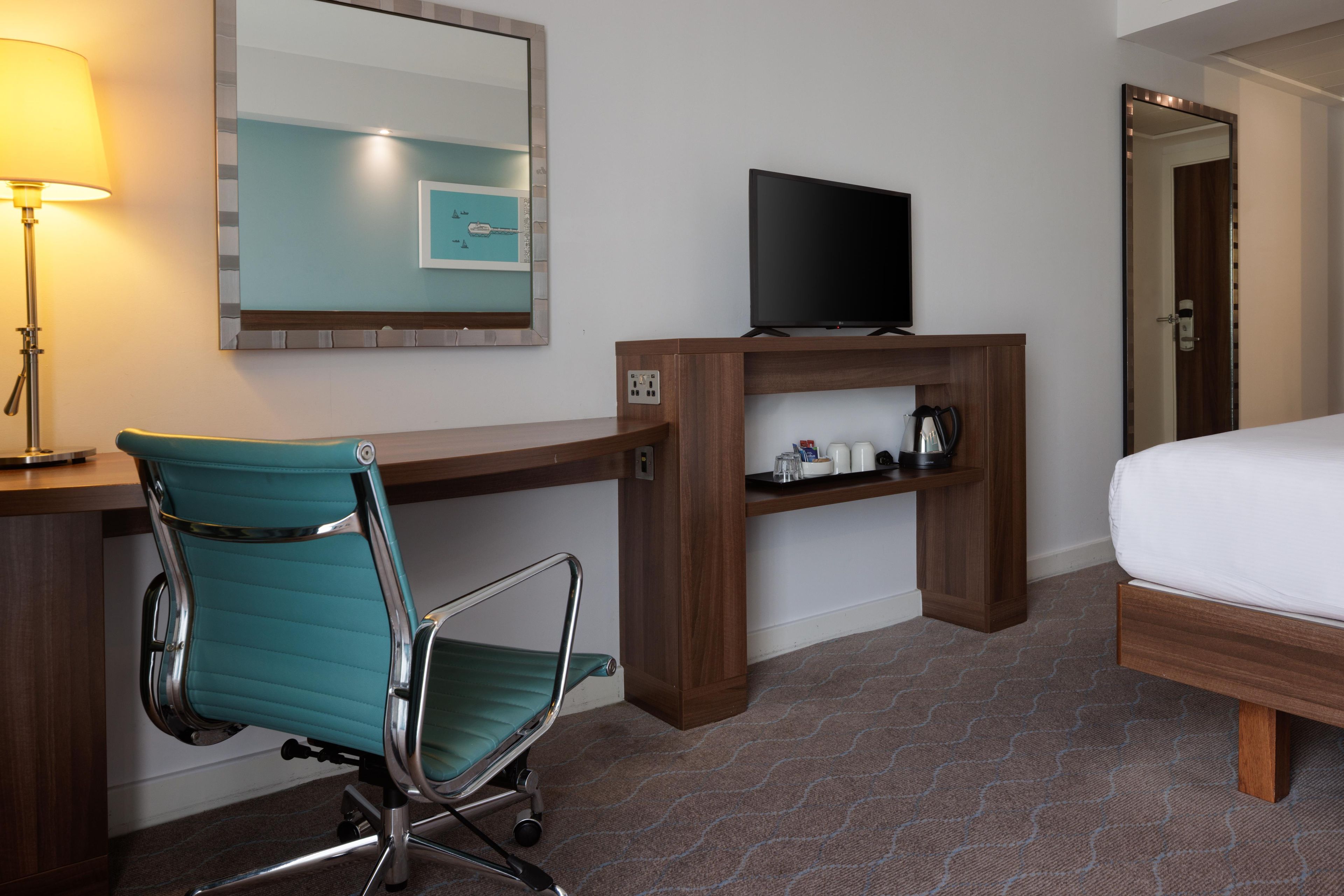 A hotel room with a desk, a chair, a bed, a television, and a mirror.