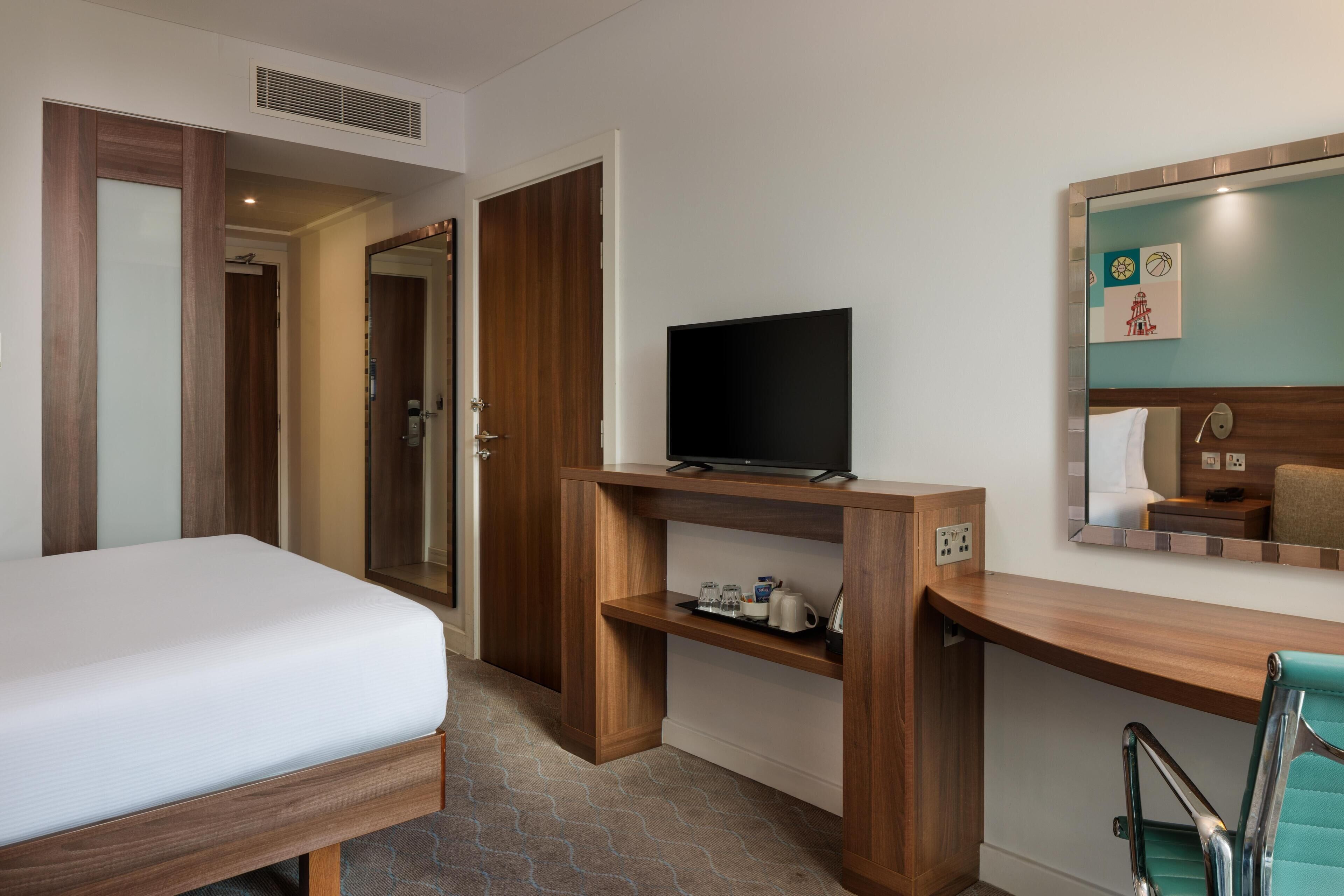 A hotel room with a bed, a TV on a wooden stand, a mirror, and a bathroom with a shower.
