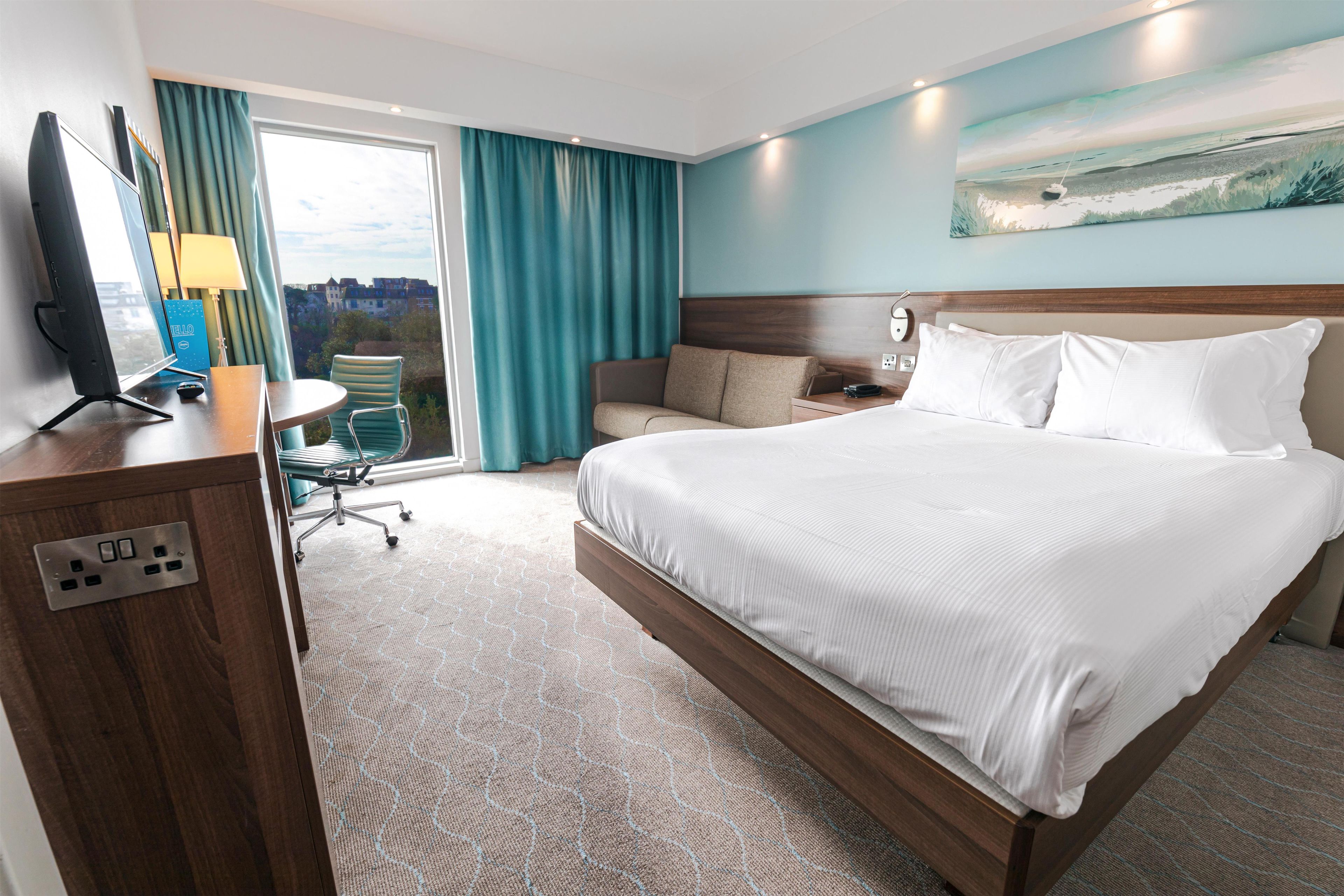 Habitación de hotel con una cama grande, un escritorio con computadora, una silla y vistas a edificios por la ventana.