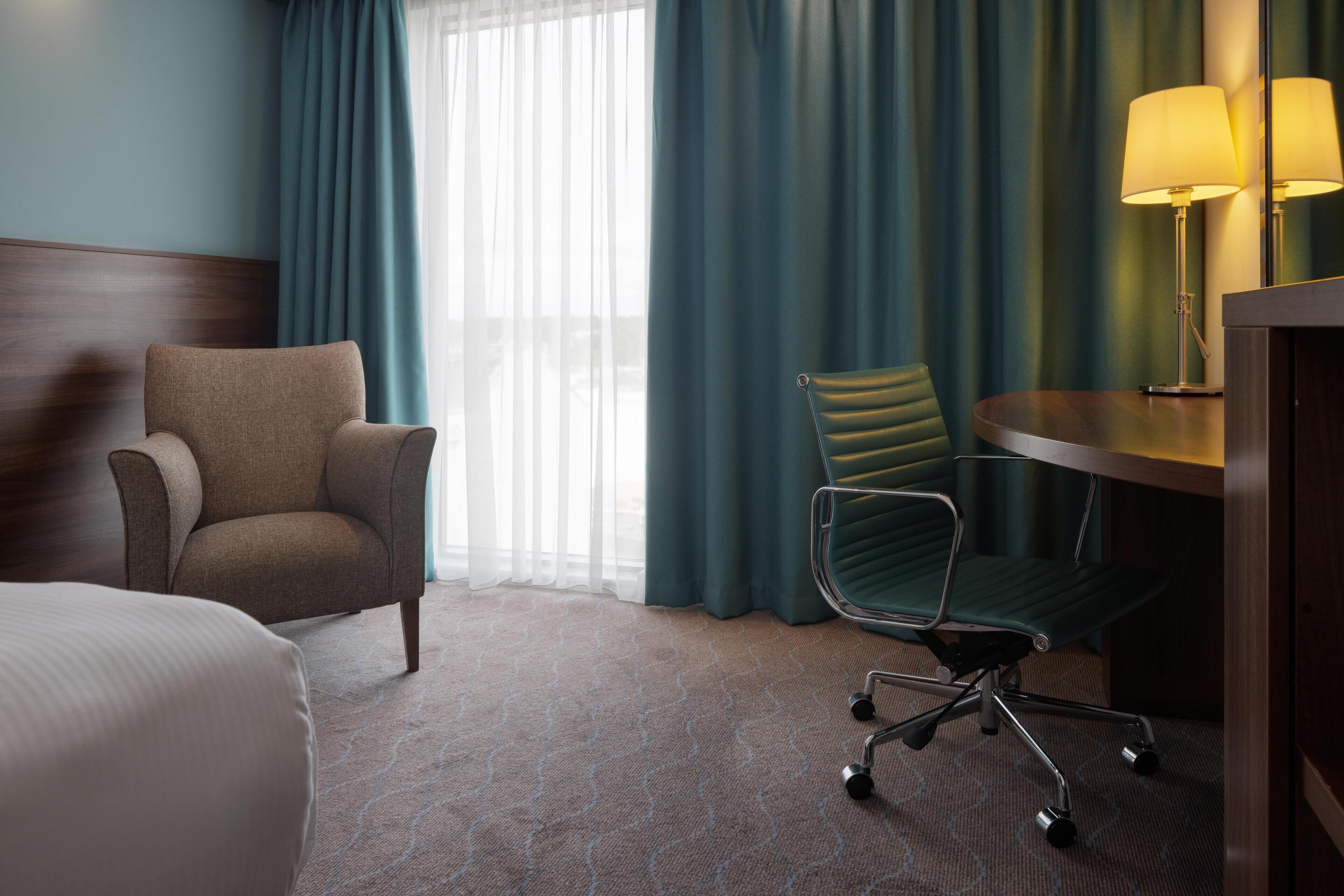 A hotel room with a bed, a chair, a desk with a lamp, and a view of the outdoors through sheer curtains.
