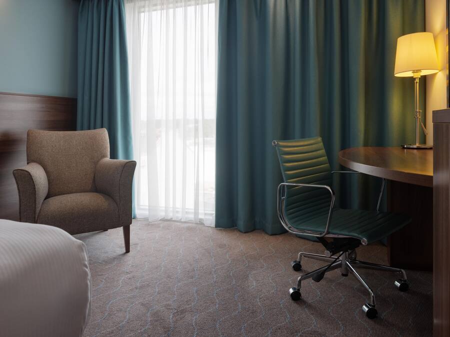 A hotel room with a bed, a desk with a lamp, a chair, and a view of the outdoors through a window with curtains.
