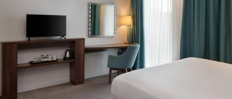 A hotel room with a bed, a desk with a television, a chair, a lamp, and a window with curtains.