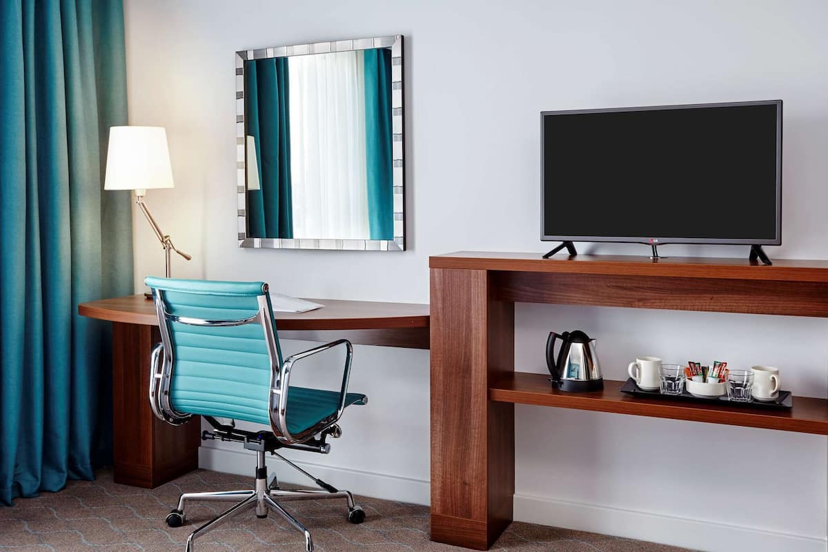 A hotel room with a wooden desk, a teal office chair, a television, a coffee maker, and a tray with cups and snacks.