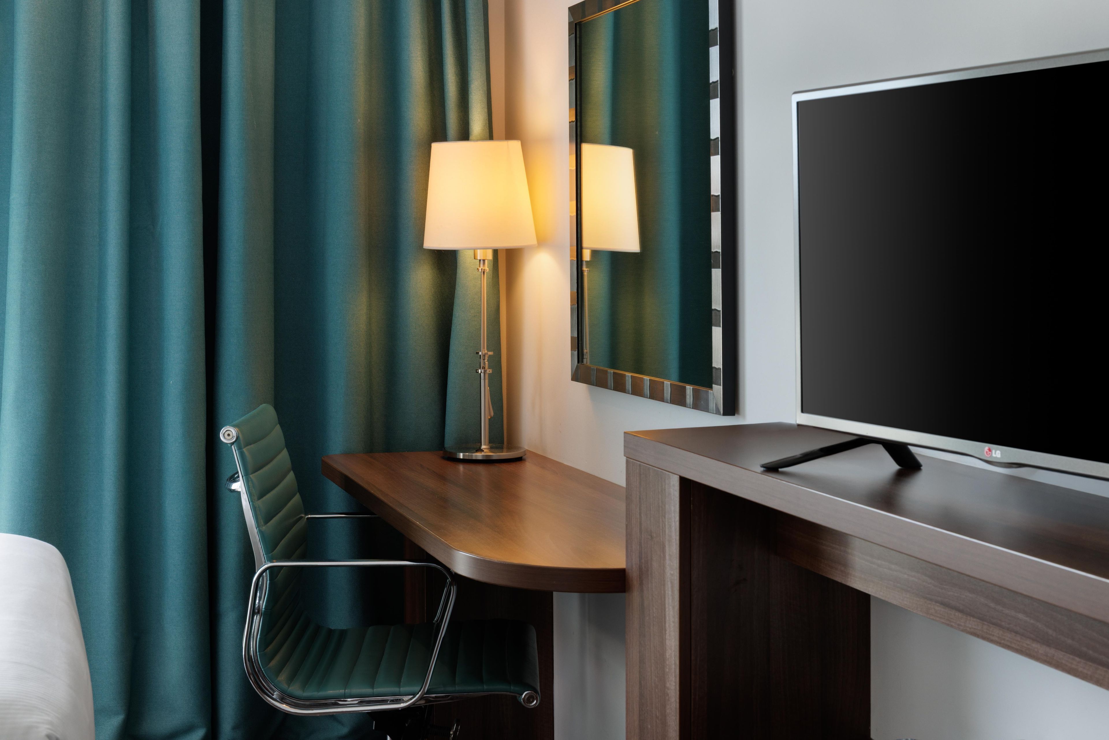 A hotel room with a bed, a desk with a lamp, a chair, a TV, and a mirror.