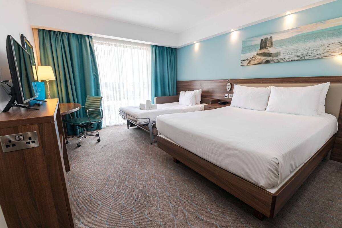 A hotel room with two beds, a desk with a computer, a chair, and a large window with curtains.
