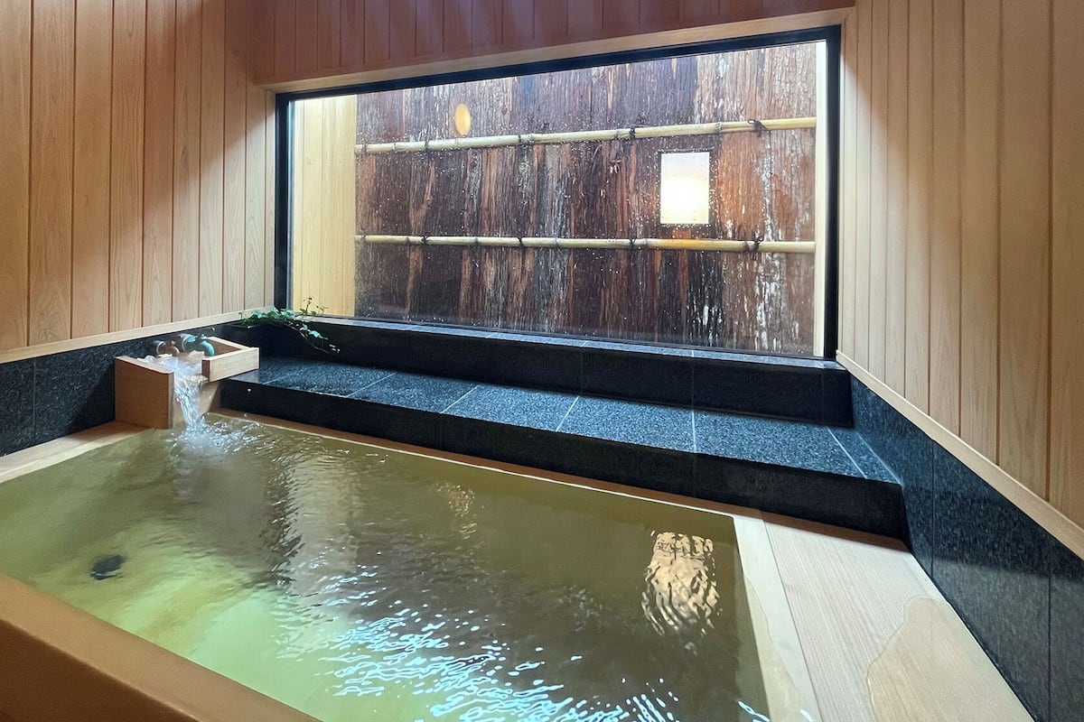 A wooden spa with a hot tub, a window with a view of a wooden structure outside.