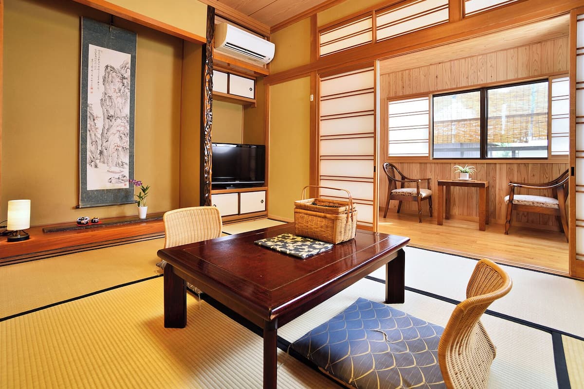 A traditional Japanese room with tatami flooring, a wooden table, chairs, a television, and a large window.