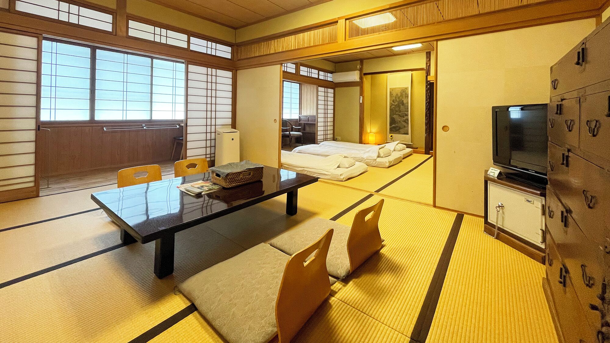 A traditional Japanese room with tatami flooring, a low table, chairs, a bed, a television, and storage cabinets.