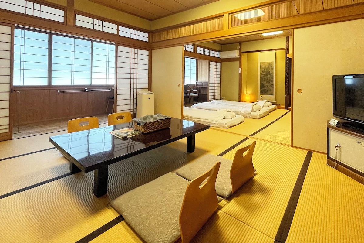 A traditional Japanese room with tatami flooring, a low table, chairs, a bed, a television, and storage cabinets.