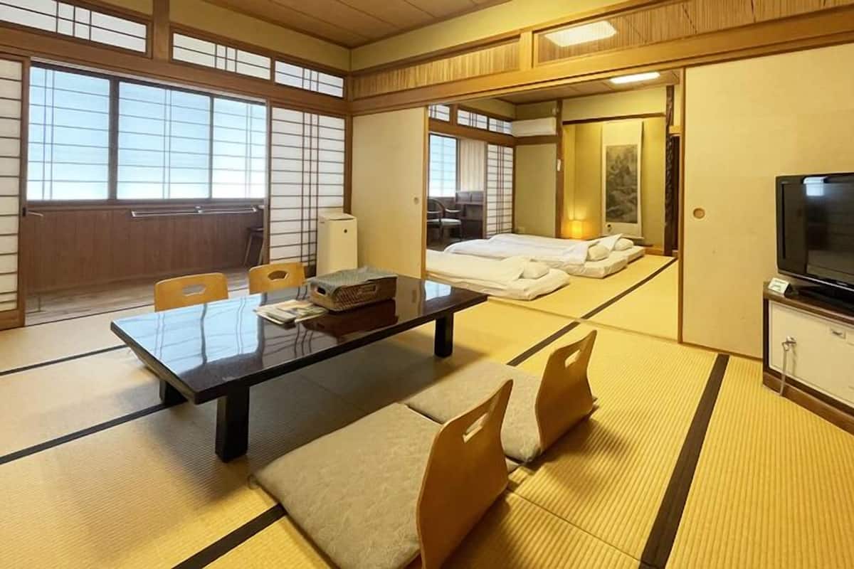 A traditional Japanese-style room with tatami flooring, a low table with chairs, a bed, a television, and a cabinet.