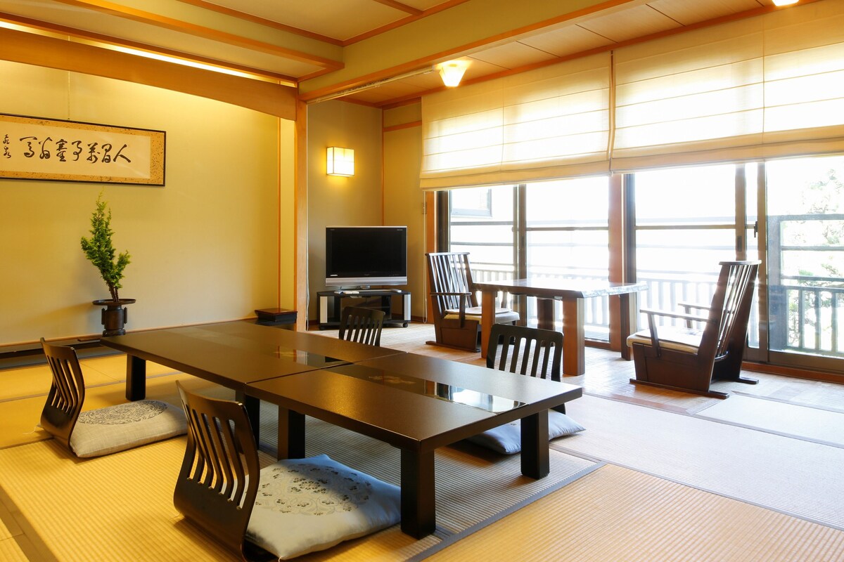 A traditional Japanese-style room with tatami flooring, a low table, chairs, a television, and a large window.
