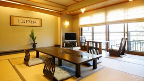 A traditional Japanese-style room with tatami flooring, a low table, chairs, a television, and a large window.