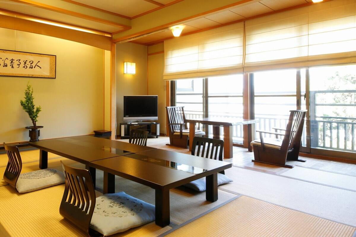 Una habitación tradicional japonesa con suelo de tatami, una mesa baja, sillas, un televisor y un ventanal grande.