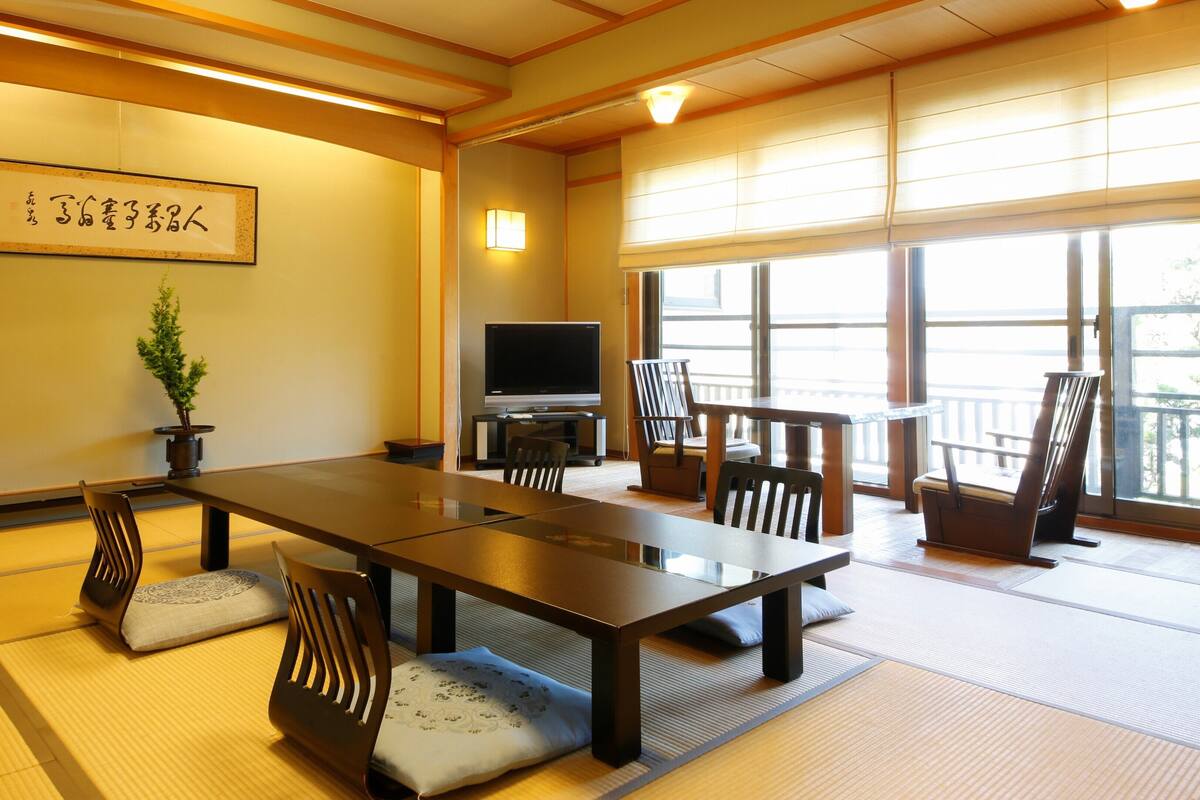 Una habitación tradicional japonesa con suelo de tatami, una mesa baja, sillas, un televisor y un ventanal grande.