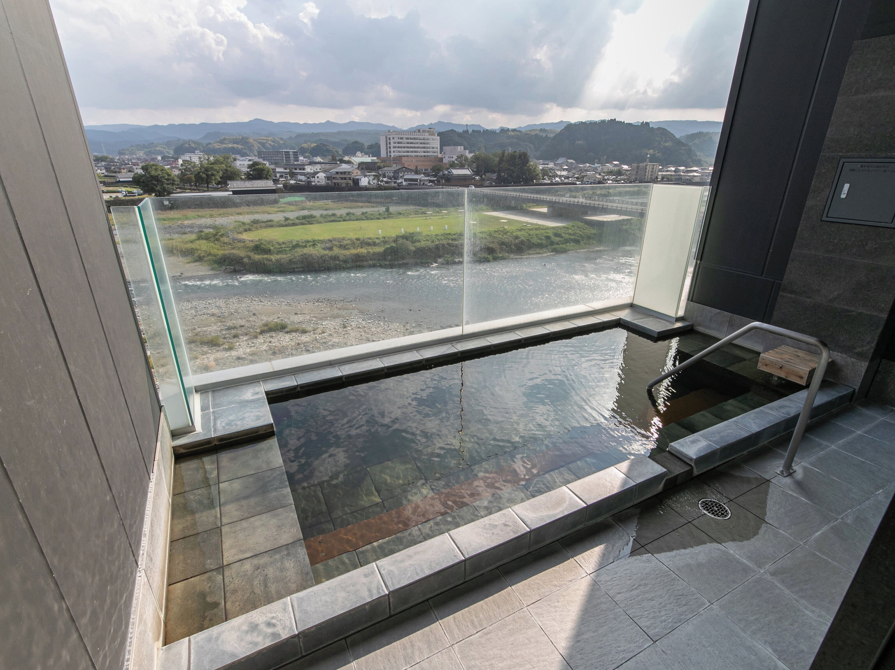 A modern indoor pool with a glass railing, a view of a river and mountains, and a stone-tiled floor.