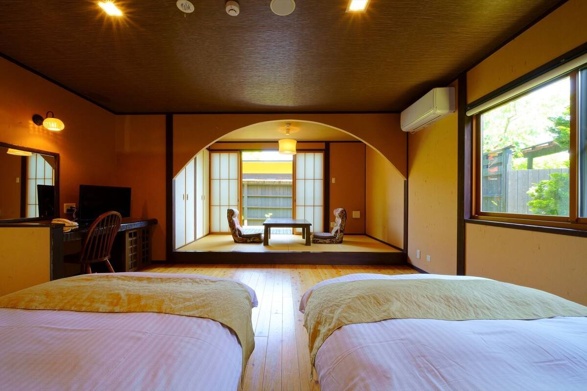 A room with two beds, a desk with a chair, a television, and a large window with a view of greenery.