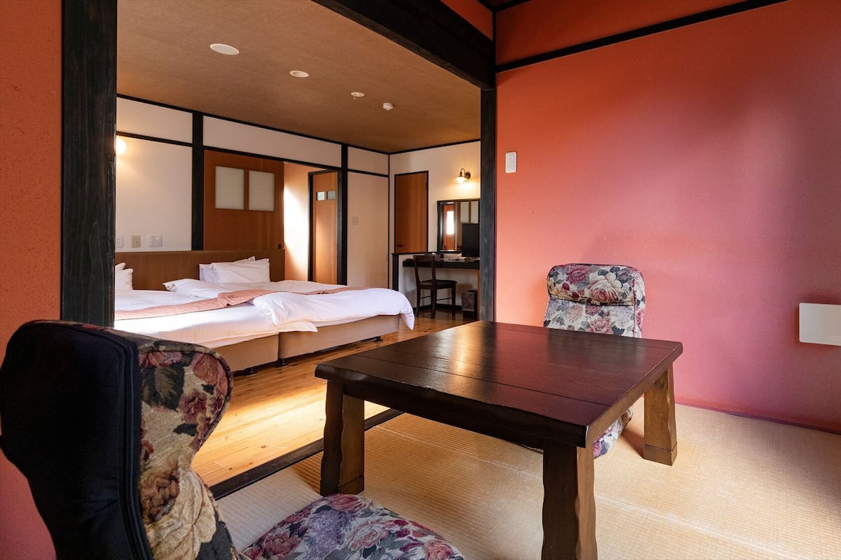 A traditional Japanese-style room with tatami flooring, a low wooden table, a bed with white bedding, and a chair with floral upholstery.