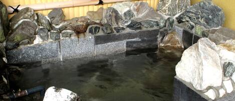 A hot spring area with a stone pool, rocks, and bamboo walls.