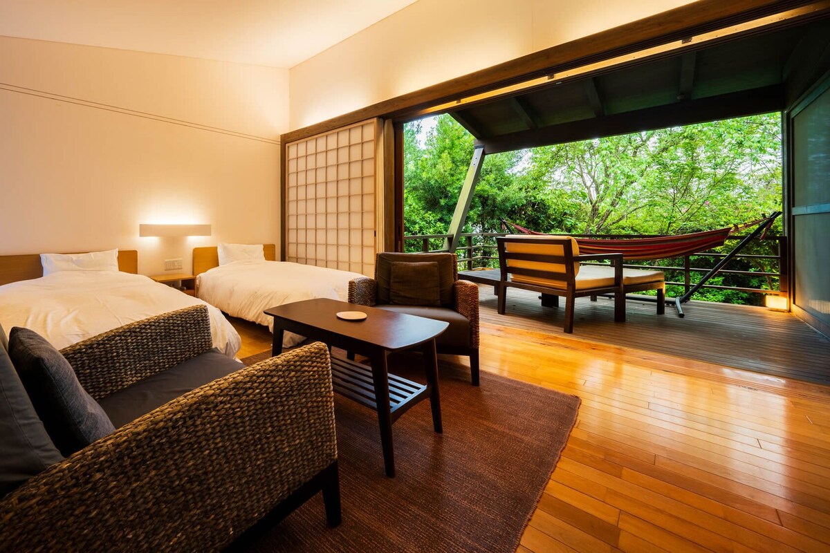 A bedroom with two beds, a sitting area with a table and chairs, and a view of greenery through a large window.