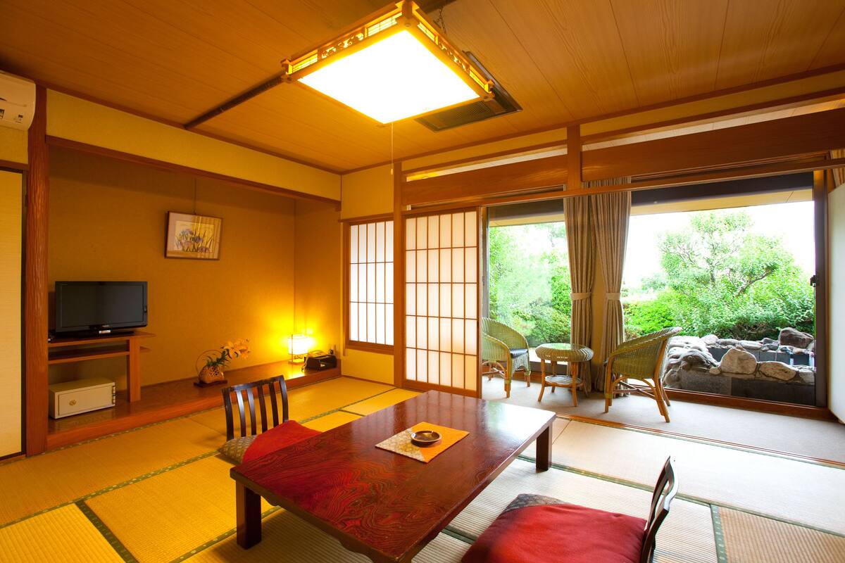 Ett traditionellt japanskt rum med tatamimattor på golvet, ett lågt bord med en röd matta, en tv på ett stativ och utsikt över grönskan genom skjutdörrar.