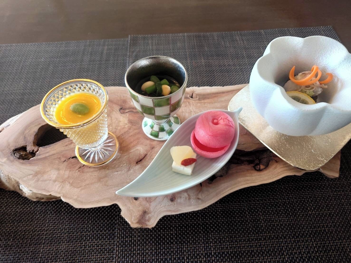 Una bandeja de madera con diversos alimentos, incluyendo un vaso con líquido amarillo, un cuenco con ingredientes verdes y marrones, un postre rosado y un plato blanco con rodajas de naranja.
