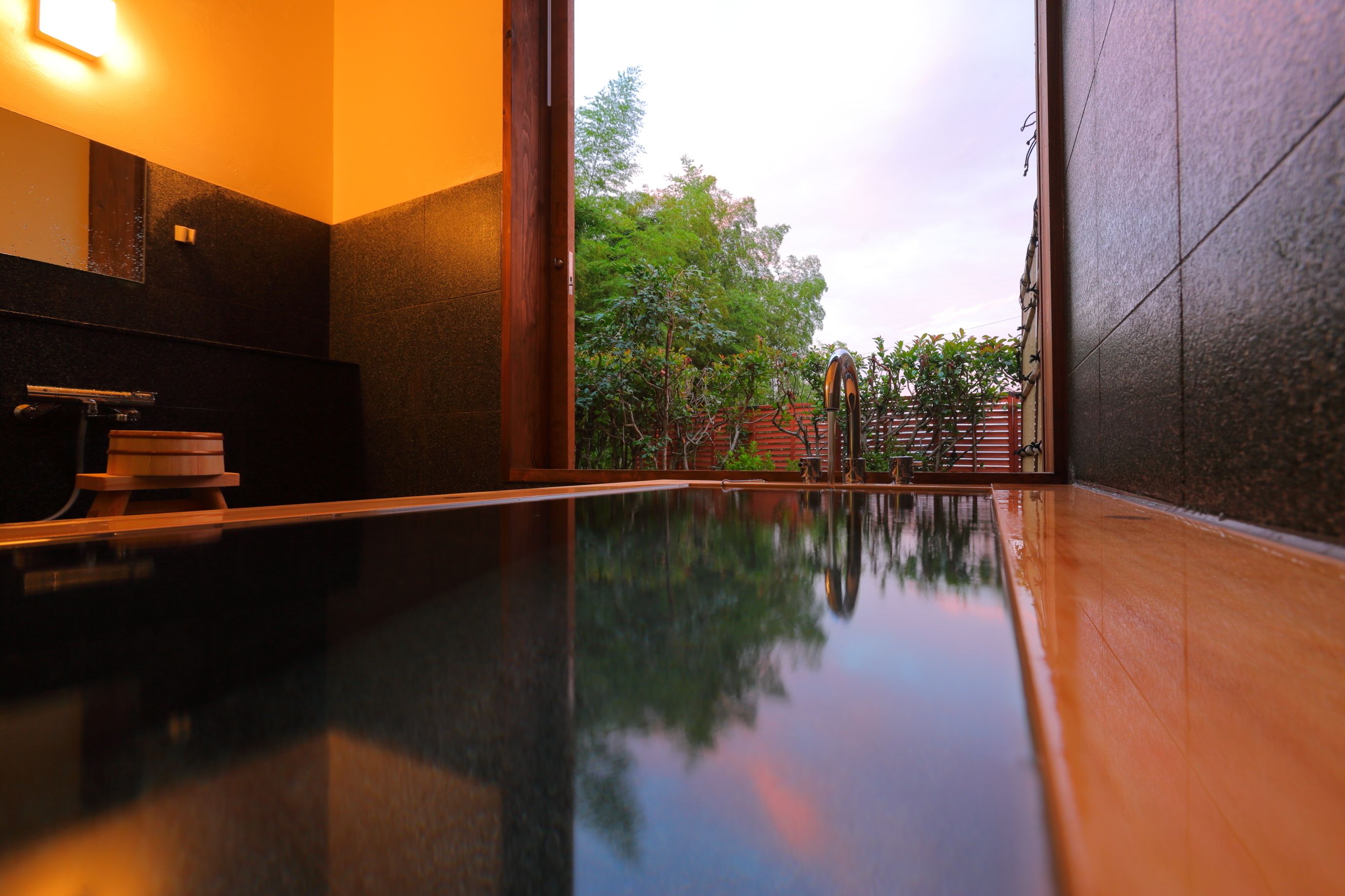 Un baño moderno con piso reflectante, un banco de madera y vistas a un jardín a través de una puerta de vidrio.