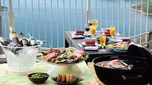 Impostazione tavola con barbecue, piatto di pesce e bevande, con vista su un corpo d'acqua.