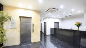 A modern reception area with a black reception desk, a potted plant, and a wall-mounted elevator. - Urban Hotel Tokyo Fuchu Nisiguchi (Fuchu)