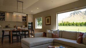 A modern kitchen with white cabinets, a dining area with a wooden table and chairs, a beige sofa with cushions, and a view of a mountainous landscape through large windows. - Craggy Range Luxury Vineyard Retreat (Tuki Tuki)
