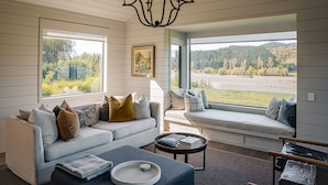 A cozy living room with a sofa, a coffee table, and a bench by the window, offering a view of a forested landscape. - Craggy Range Luxury Vineyard Retreat (Tuki Tuki)