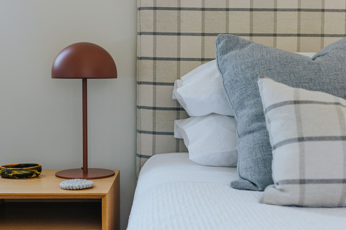 A neatly made bed with a plaid headboard, a round table with a lamp, and a small bowl on the nightstand.
