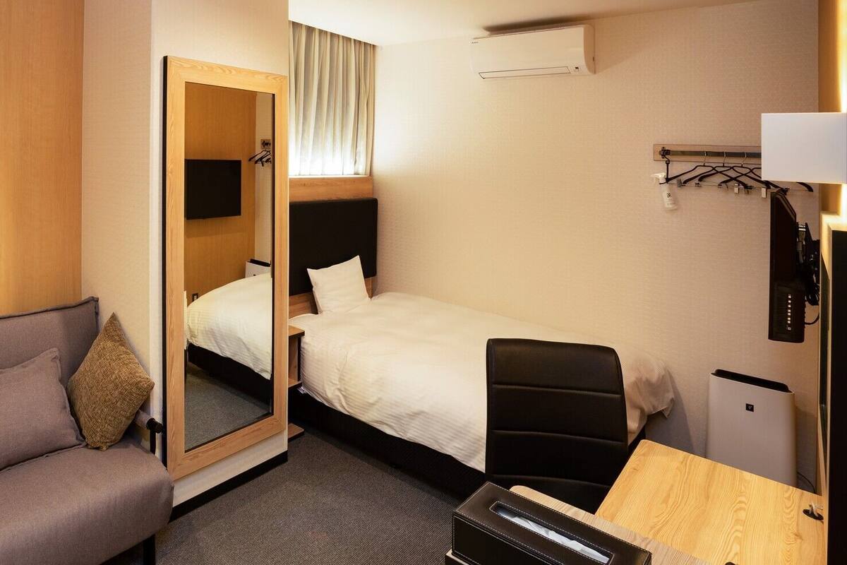 A hotel room with a bed, a sofa, a desk, a chair, a mirror, and an air conditioning unit.