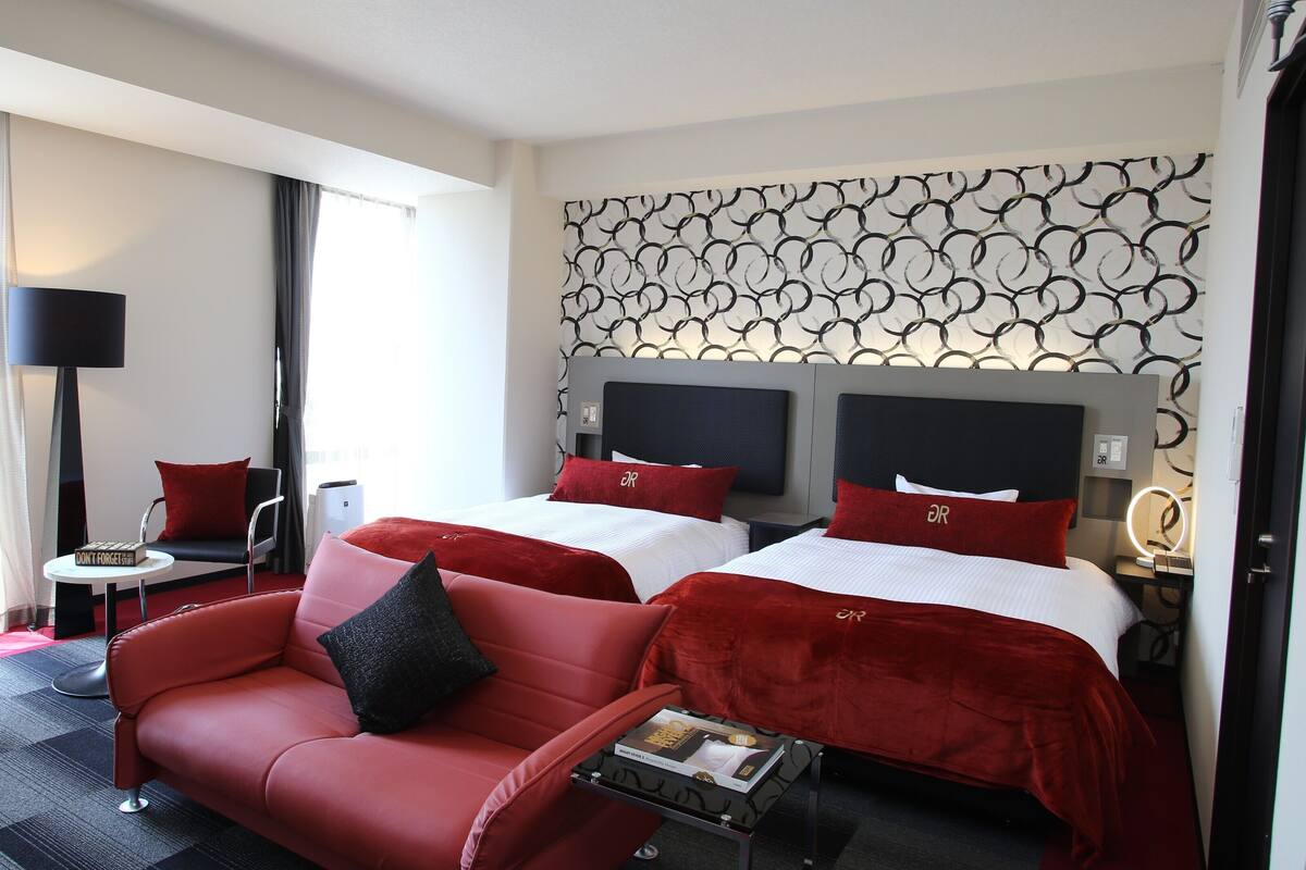 A hotel room with two beds, a red sofa, a small table, a lamp, and a patterned wall.