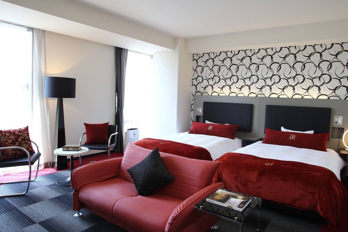 A hotel room with a red sofa, two beds with matching red pillows, a patterned wall, and a modern lamp.