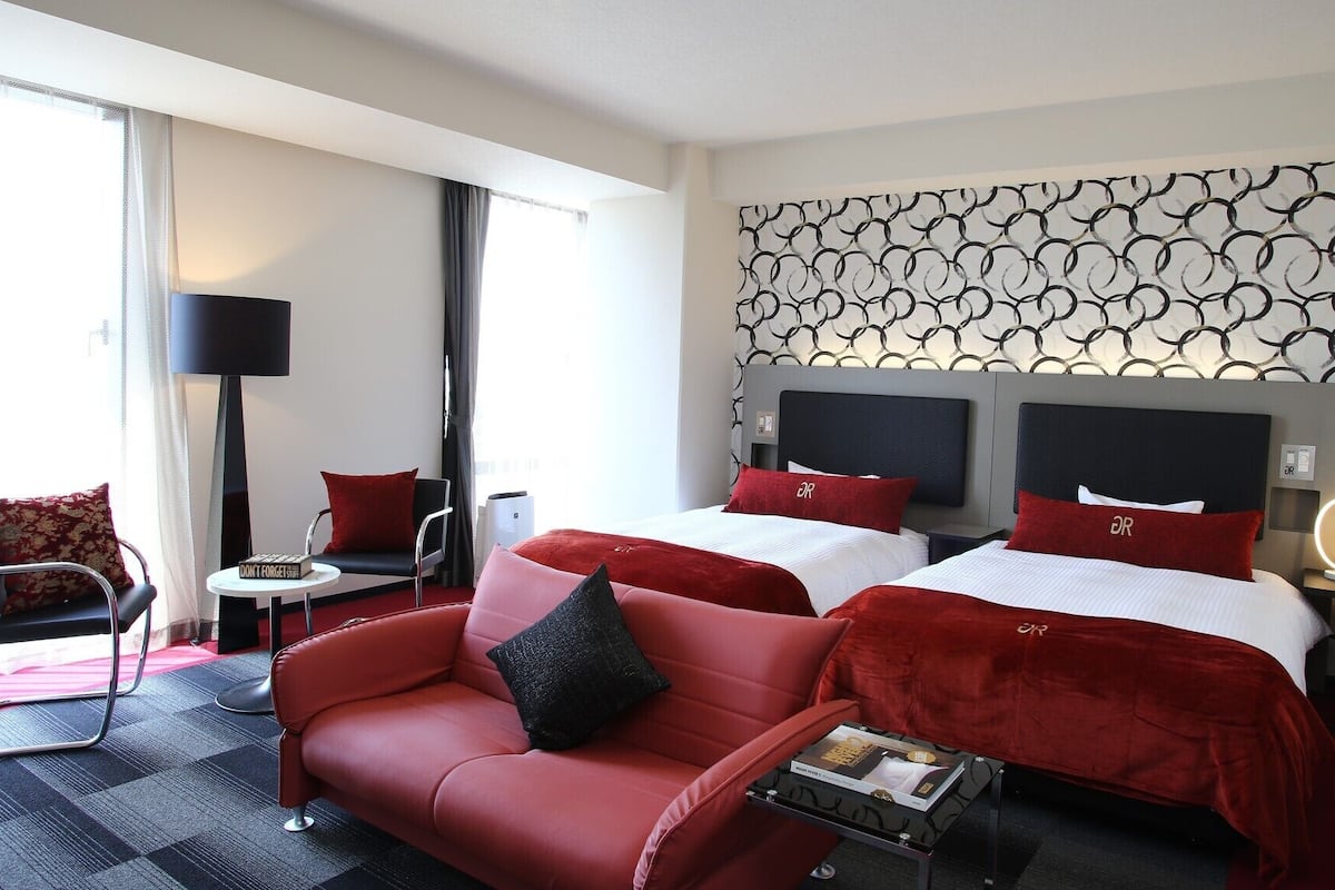 Habitación de hotel con sofá rojo, dos camas con almohadas rojas a juego, pared estampada y una lámpara moderna.