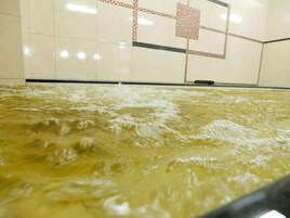 Una habitación con una amplia fuente de agua poco profunda en el centro, paredes cubiertas de mosaicos con un diseño geométrico y un objeto negro en el suelo.