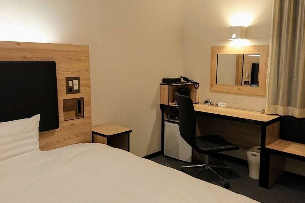 A hotel room with a bed, a desk, a chair, a mirror, and a small trash bin.
