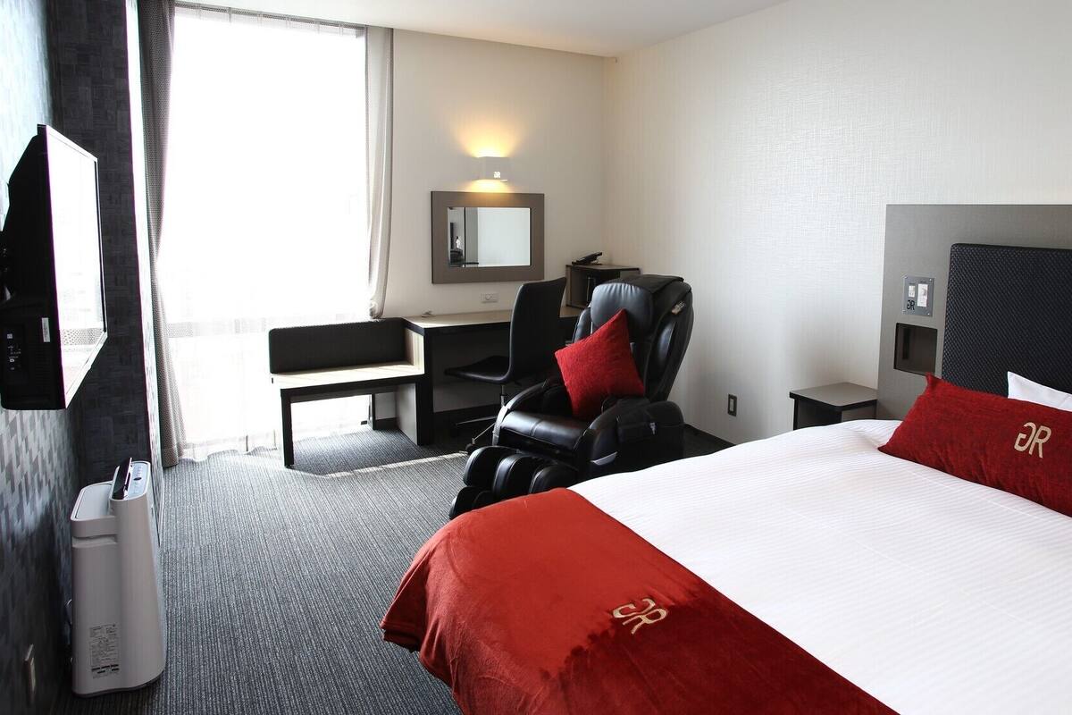 A hotel room with a bed, a massage chair, a desk, a chair, a TV, and a mirror.