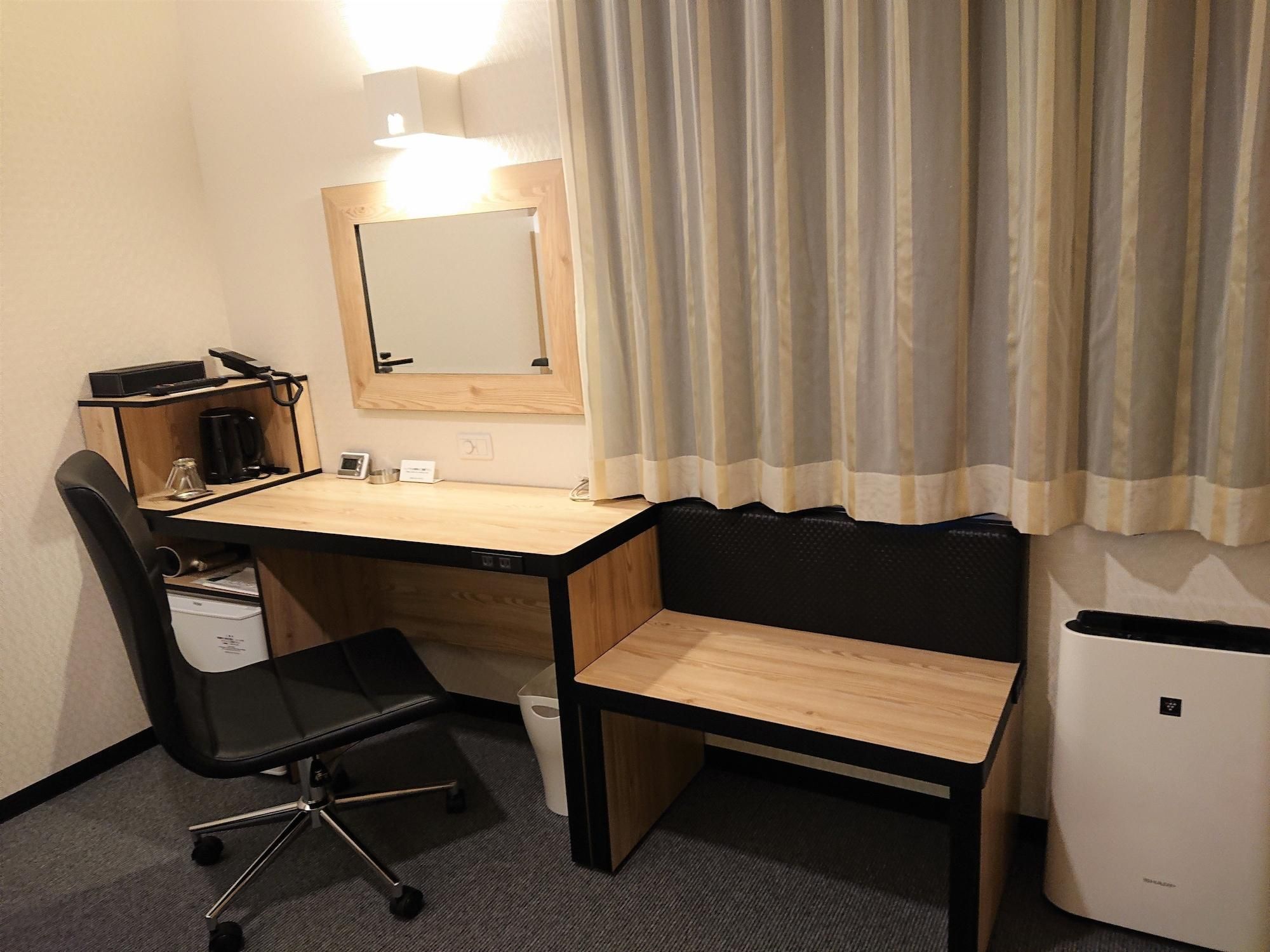 A compact hotel room with a wooden desk, a black office chair, a small waste bin, a white mini-fridge, and a mirror above the desk.