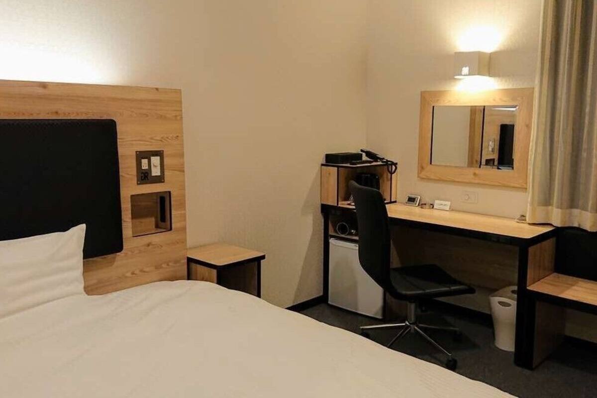 A hotel room with a bed, a desk, a chair, a mirror, and a small trash bin.