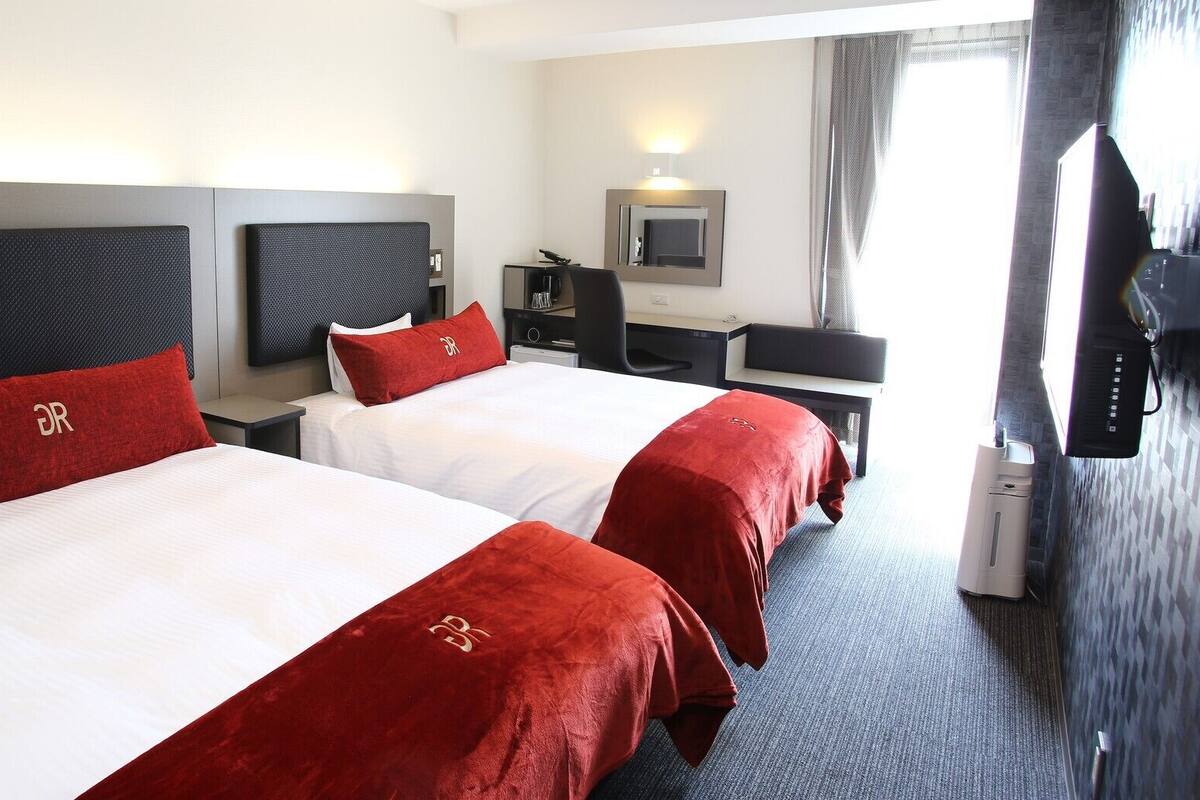 A hotel room with two beds, a desk, a chair, a TV, and a window.