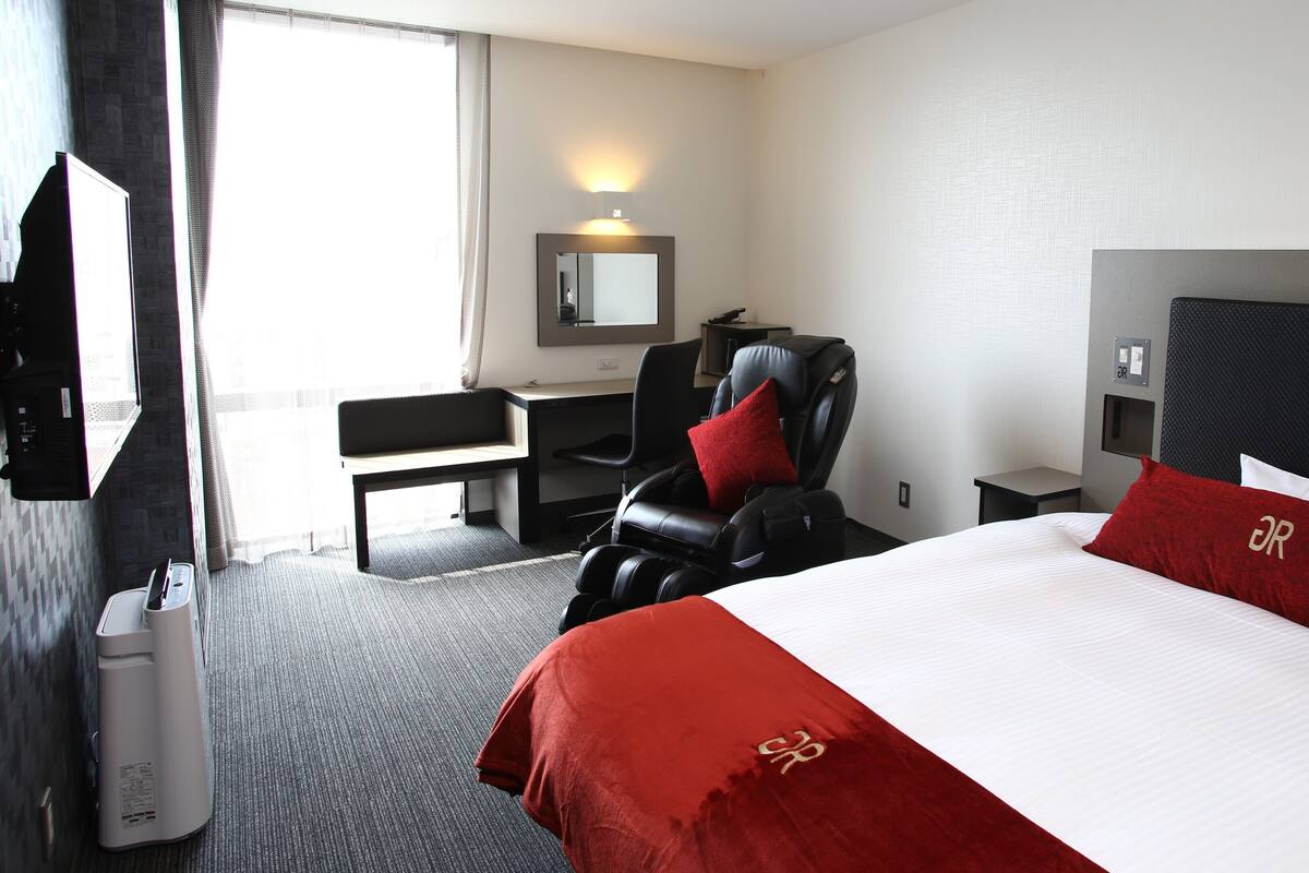 A hotel room with a bed, a massage chair, a desk, a chair, a TV, and a mirror.